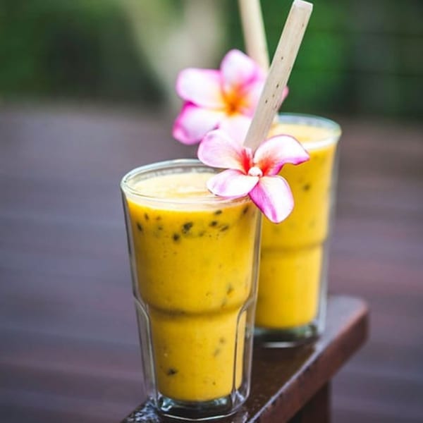 Two vibrant yellow passion fruit smoothies, garnished with pink flowers and wooden straws, on a wooden surface.